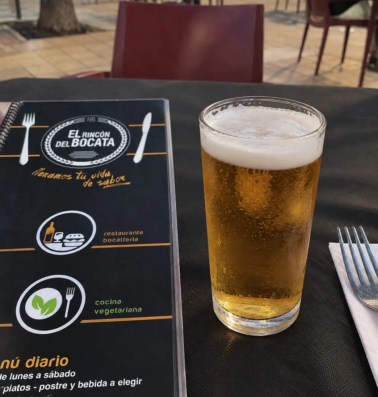 A glass of draught beer with a head of foam on a terrace table, beside the restaurant menu open at its cover.
