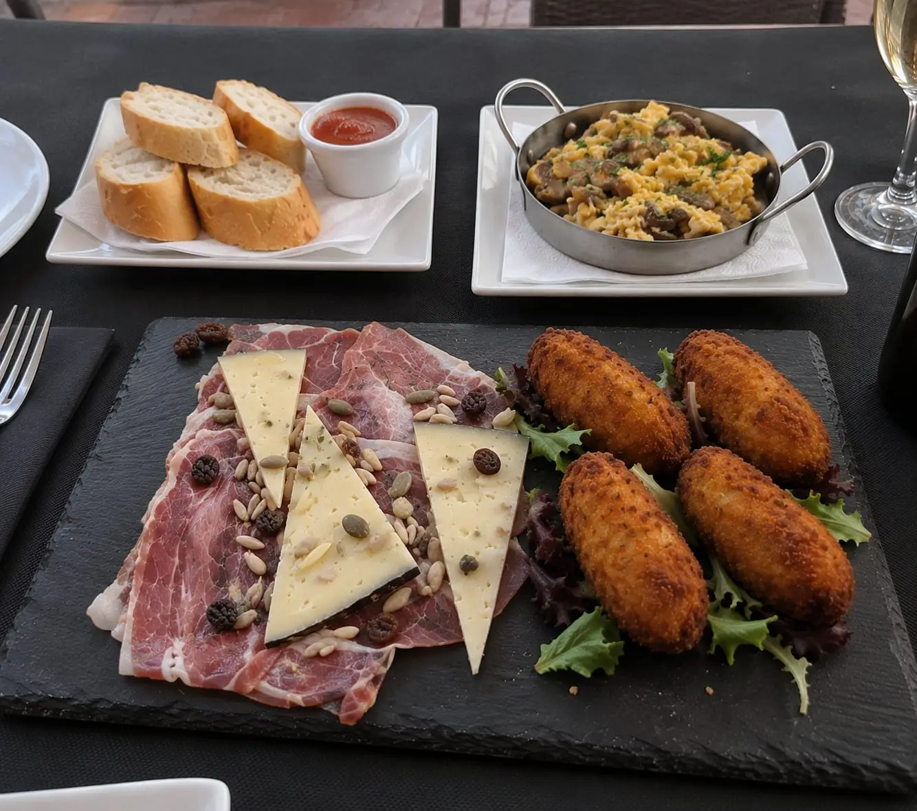 Mesa de entrantes para compartir: tabla de jamón y queso con croquetas, una sartén de revuelto con setas y una cesta de pan con salsa.