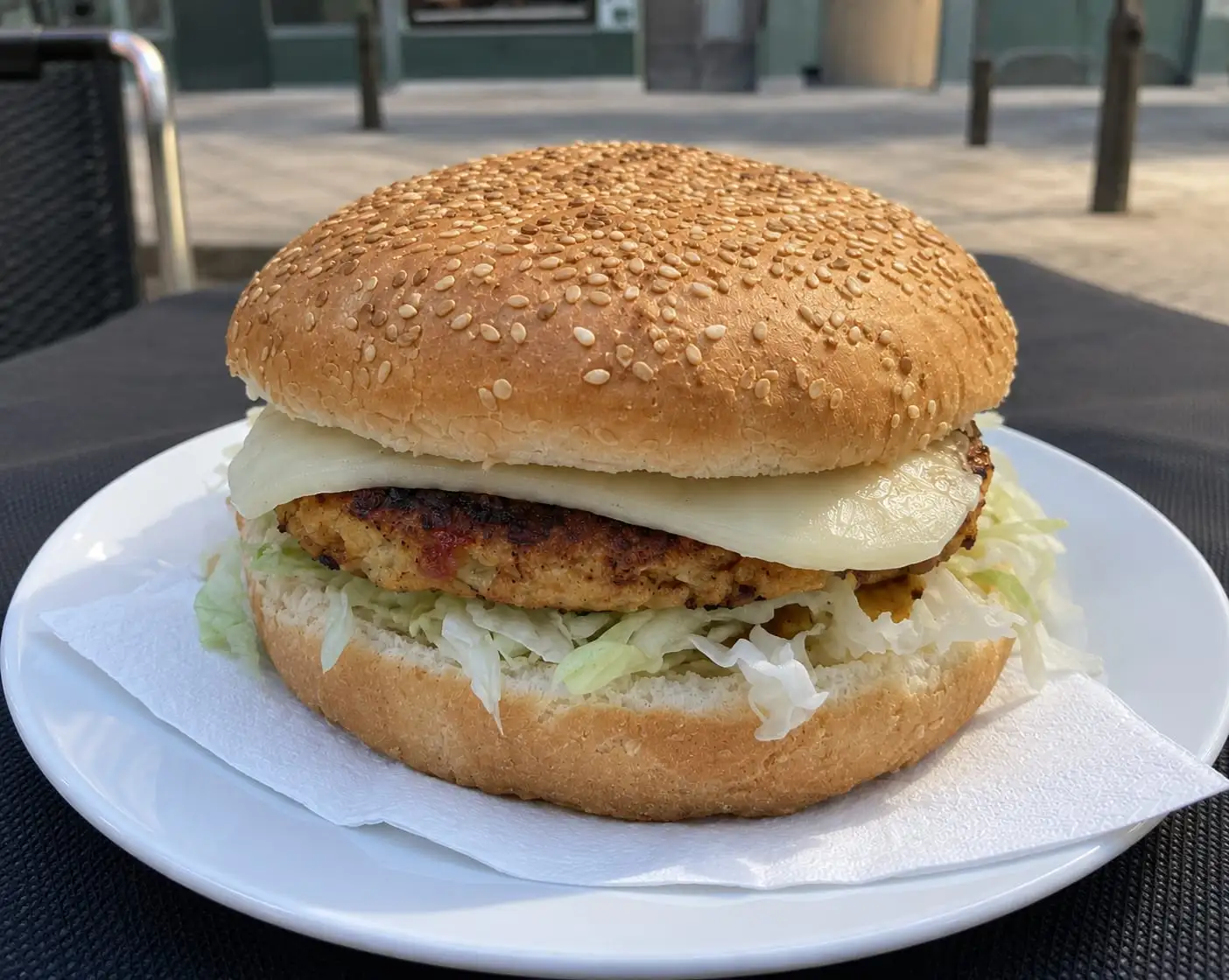 A burger in a sesame bun with melted cheese and lettuce, served on a white plate on the terrace.