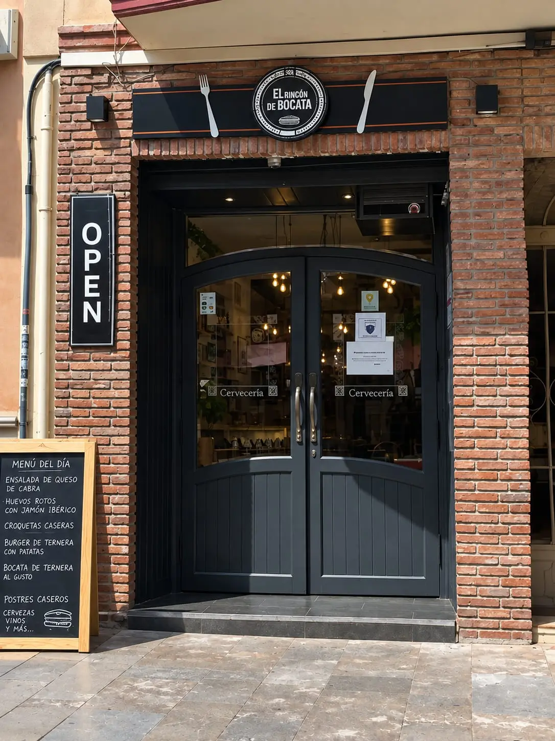 Entrada d'El Rincón del Bocata: porta doble fosca entre pilars de rajola, rètol amb coberts i pissarra amb el menú del dia a la vorera.