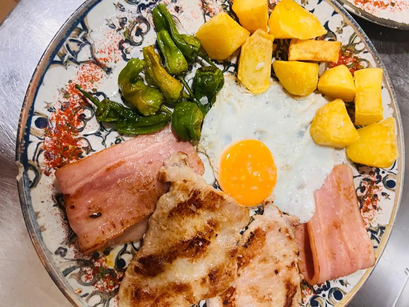 A plate with a fried egg, potatoes, ham and grilled pork loin, with Padrón peppers.