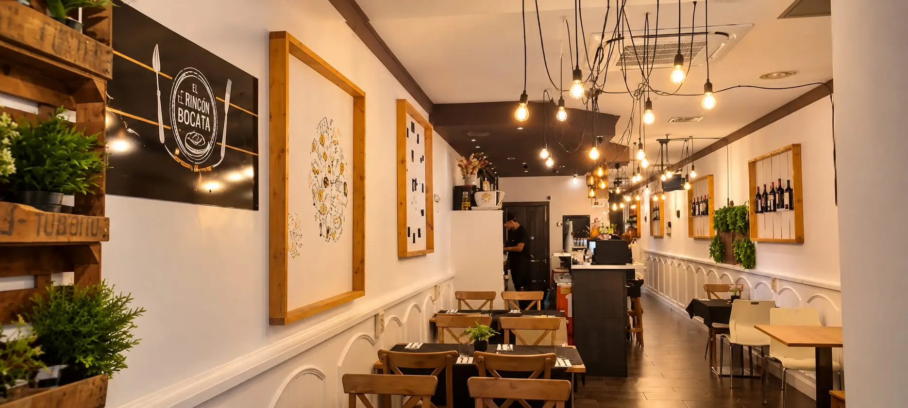 La sala allargada del restaurant: taules de fusta a un costat, la barra al fons i una filera de bombetes penjant del sostre.