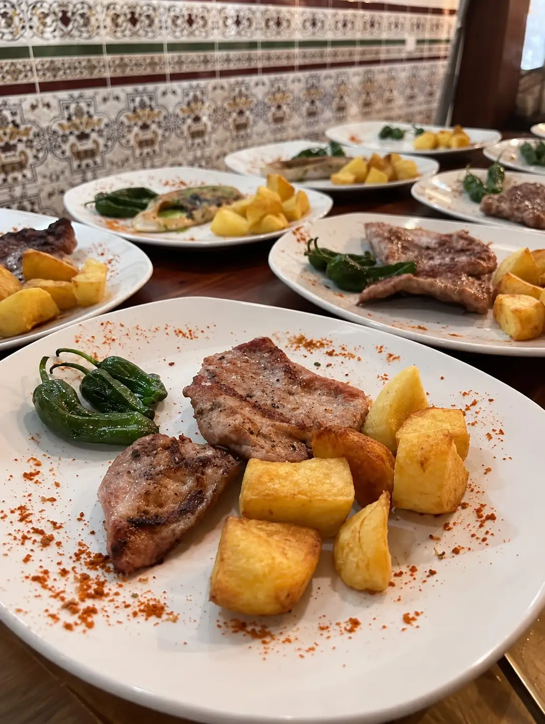 Several plates lined up on the table: grilled meat with potatoes and green peppers, in front of the dining room tiled wall.