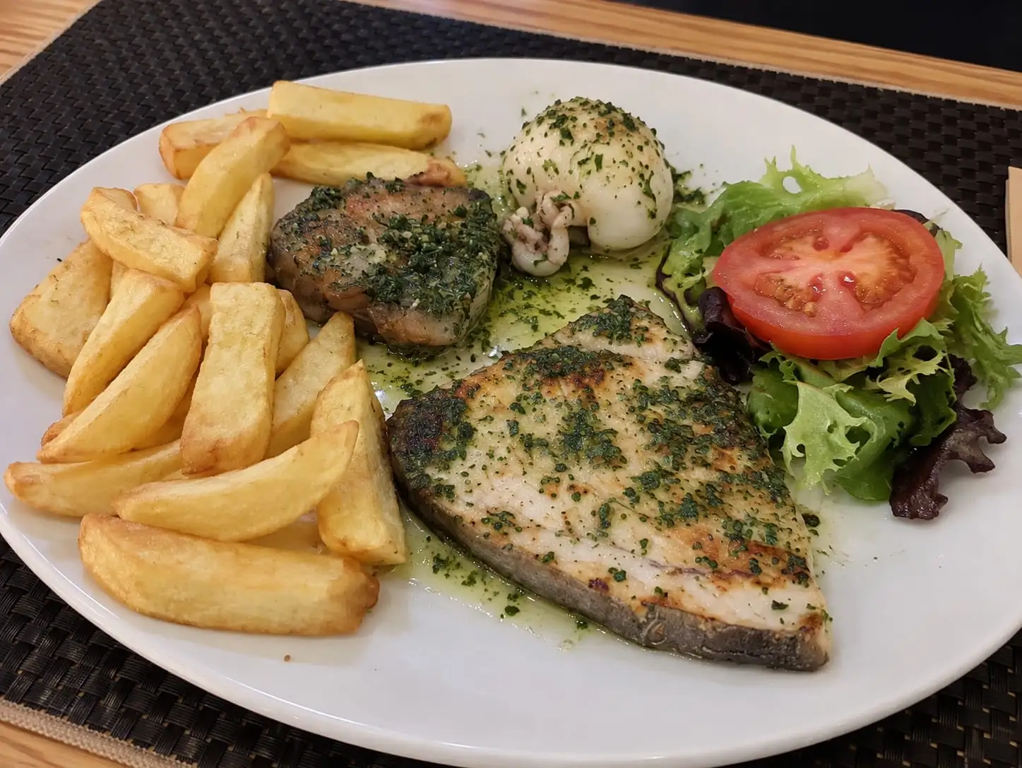 Plate of grilled fish with green sauce, a stuffed squid, chips and salad with tomato.