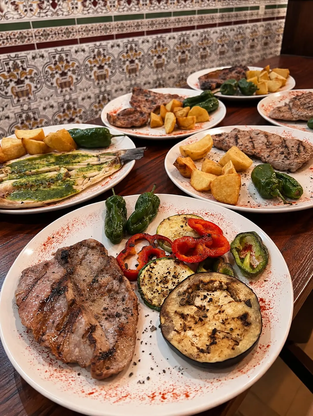 Taula amb diversos plats: bistec a la planxa amb verdures rostides, i al fons una plata amb peix i salsa verda.
