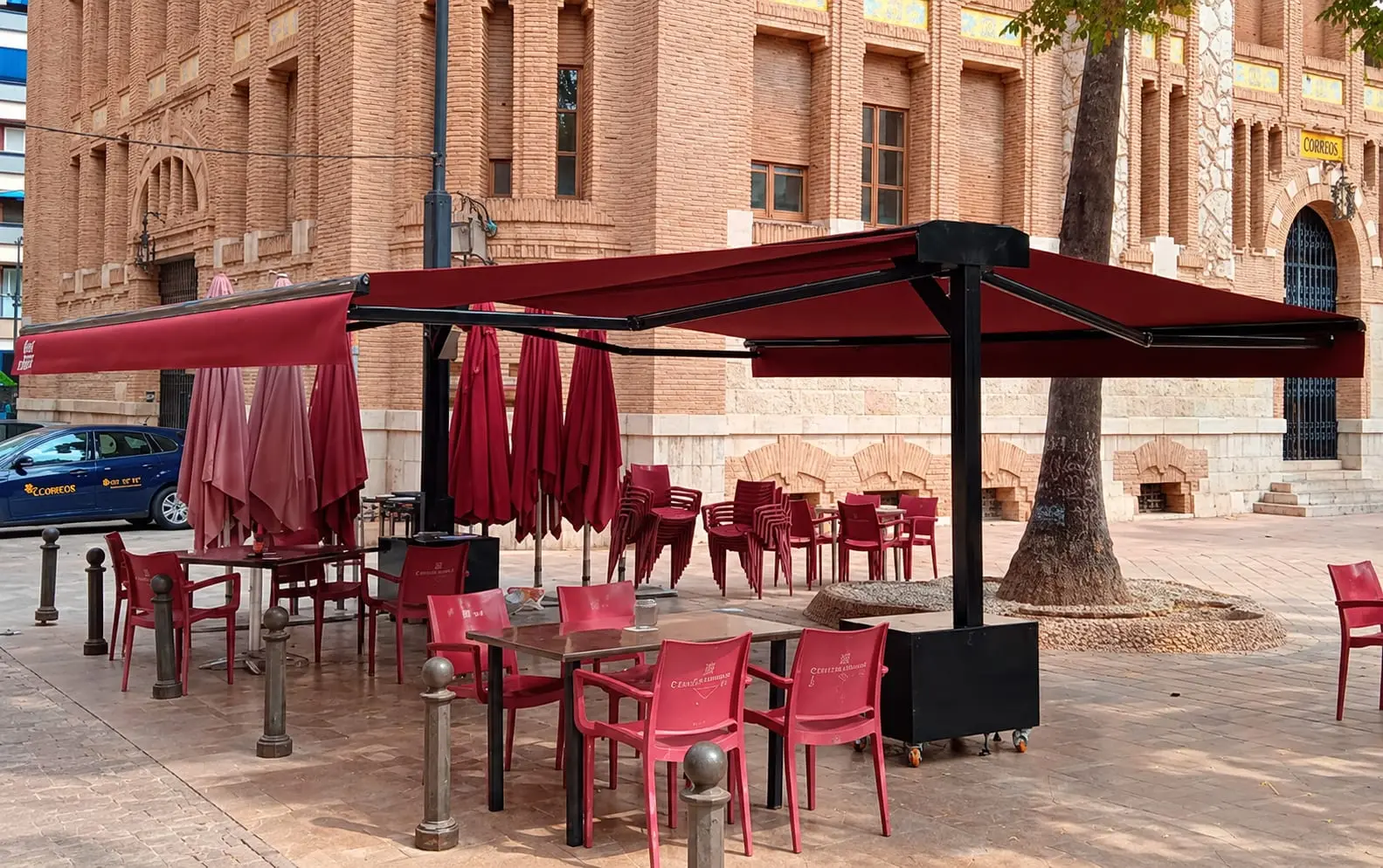 La terrassa a la plaça Tetuan: taules i cadires granats sota dos tendals, amb l'edifici de Correus darrere.