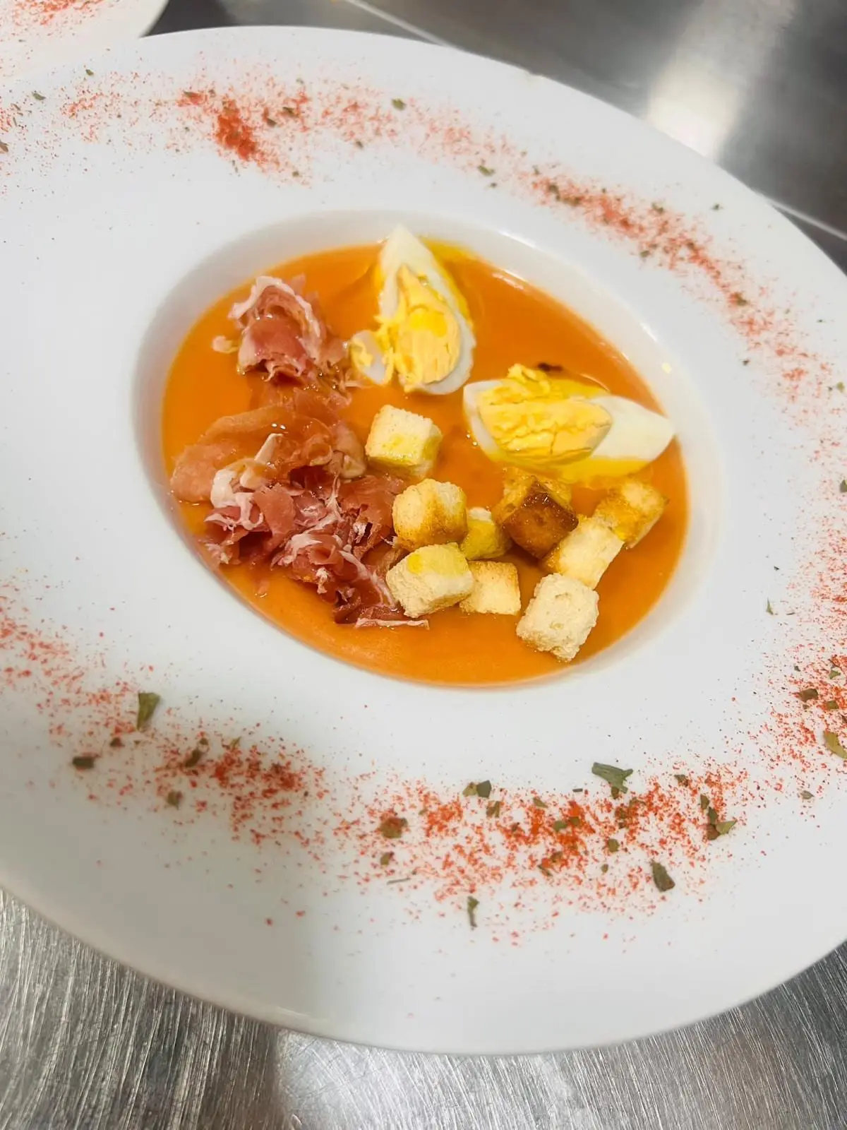 Salmorejo en plato hondo con huevo duro picado, virutas de jamón serrano y picatostes, con pimentón espolvoreado en el borde.