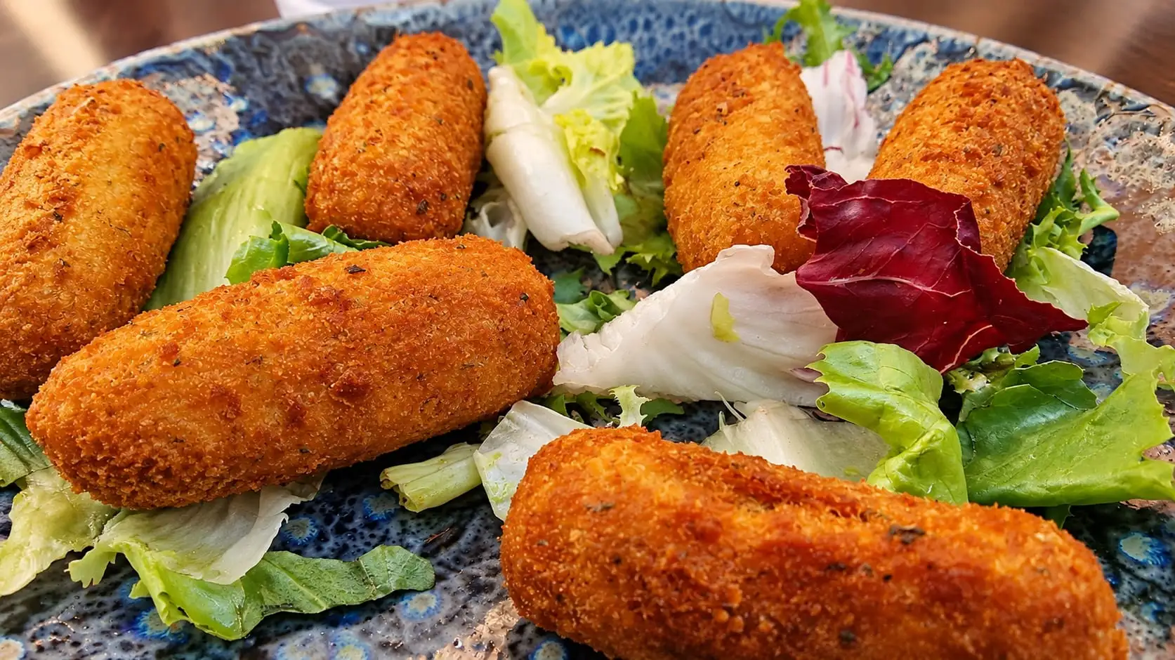 Six freshly fried croquettes on a blue plate, with salad leaves between them.