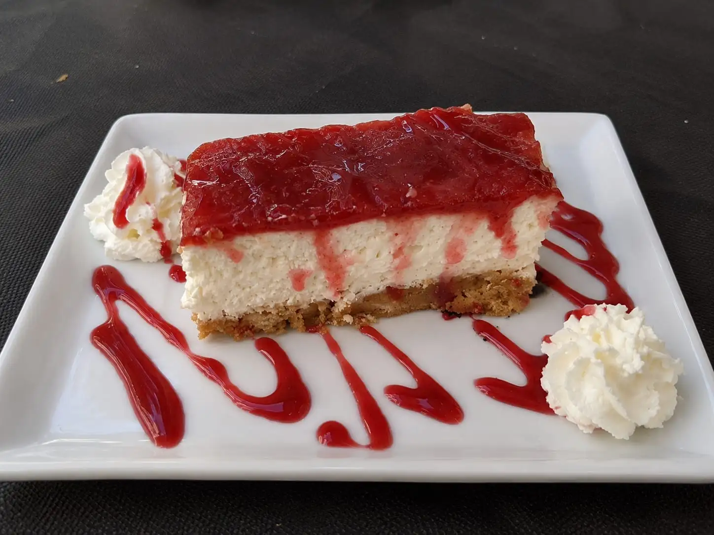 Slice of cheesecake topped with strawberry jam, whipped cream at the sides and a drizzle of coulis on the plate.