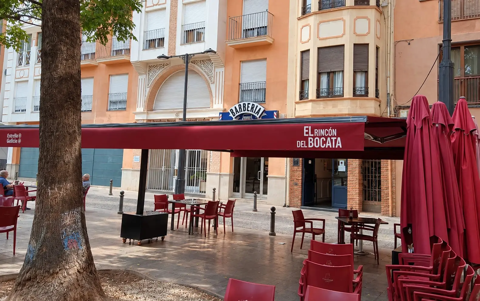 La terrassa vista des de la plaça: el tendal granat amb el nom del restaurant damunt les taules i les cadires roges, a l'ombra d'un arbre.