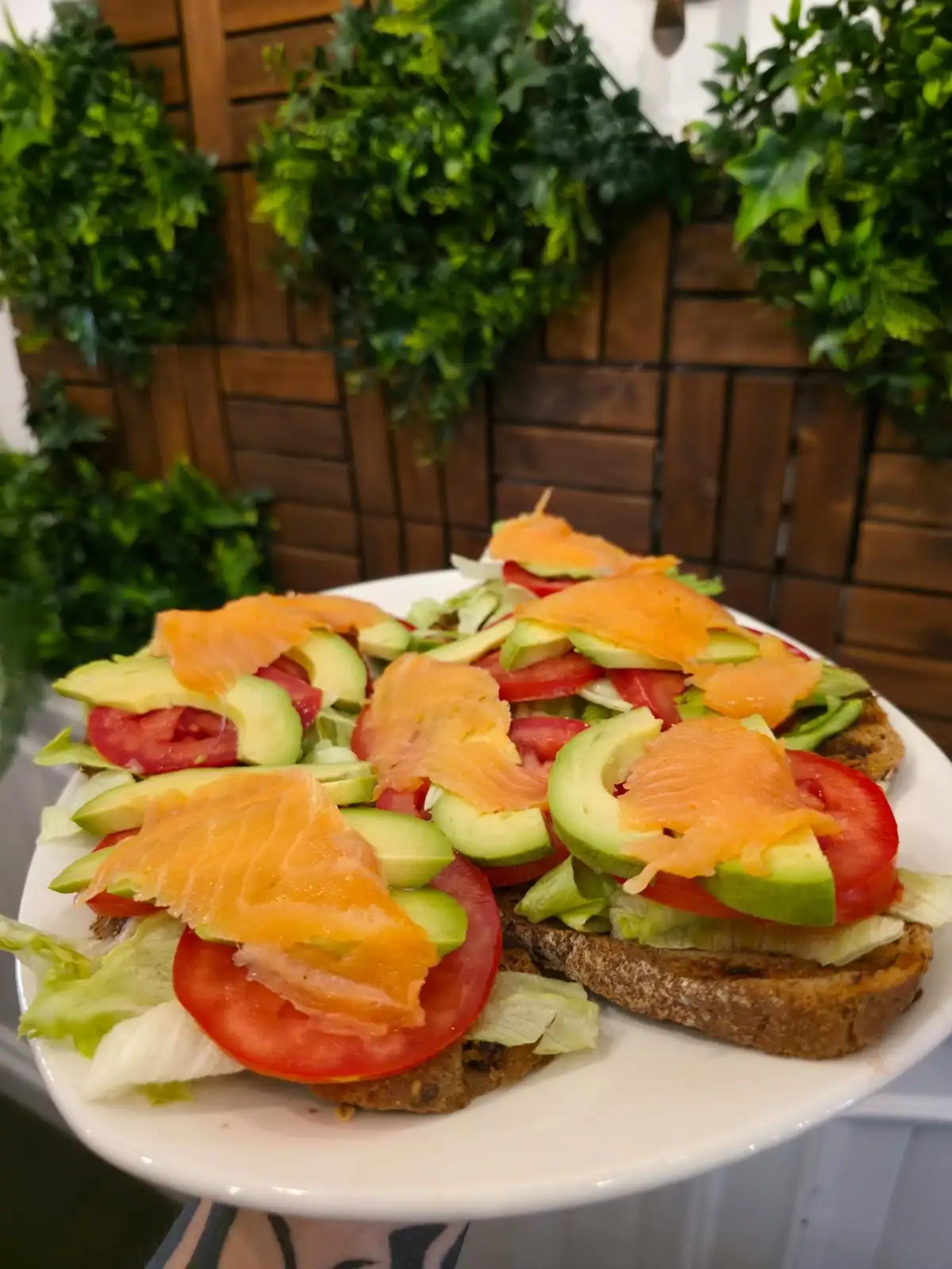 Two slices of seeded toast with lettuce, tomato, sliced avocado and smoked salmon, served on a white plate.